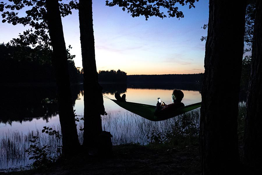 Client Center - Man Uses a Phone While Laying in a Hammock by a Lake at Dusk